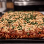A full tray of baked ziti just out of the oven, topped with golden cheese and fresh chopped herbs.